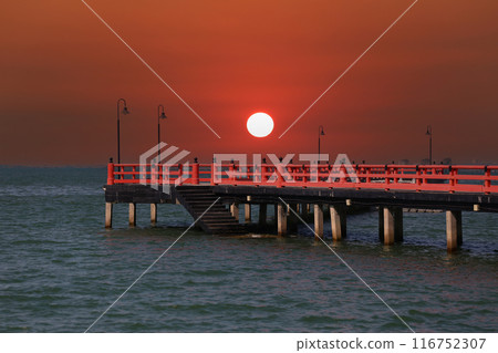 Red bridge that extends into the sea and view of the sunset in the evening time. 116752307