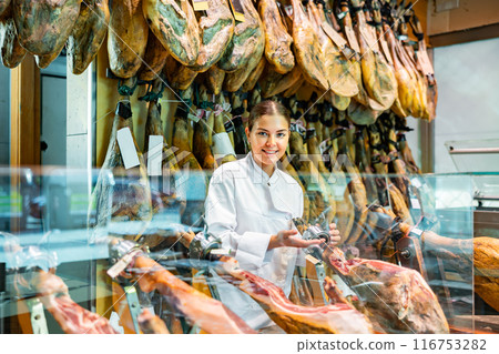 Young salesgirl in white uniform offering Iberian jamon in butcher shop Young salesgirl in white uniform offering Iberian jamon in butcher shop 116753282