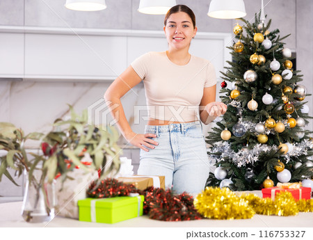 Joyful young woman in casual clothes holding wrapped gift next to Christmas tree in living room at home Joyful young woman in casual clothes holding wrapped gift next to Christmas tree in living room at home 116753327