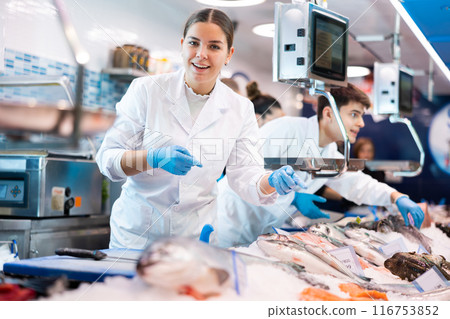 Friendly woman seller in a white coat and gloves invites you to buy fresh fish in supermarket Friendly woman seller in a white coat and gloves invites you to buy fresh fish in supermarket 116753852