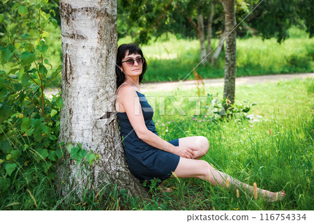 50 years old russian woman in short dress and sunglasses sitting and resting under tree shadow on the summer day  50 years old russian woman in short dress and sunglasses sitting and resting under tree shadow on the summer day  116754334