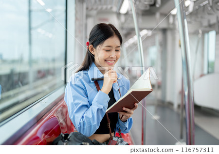 A happy Asian woman is writing her diary while commuting in the city with a sky train. A happy Asian woman is writing her diary while commuting in the city with a sky train. 116757311