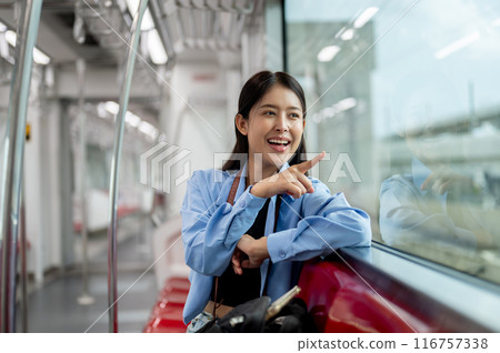 A cheerful Asian woman is enjoying the view outside while sitting on a seat in a sky train. 116757338