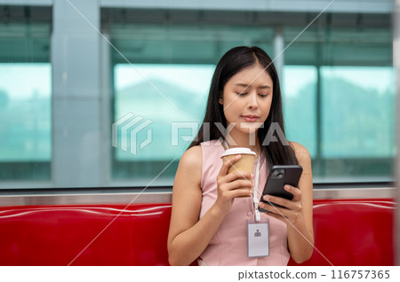 Thoughtful Asian businesswoman is sipping coffee and reading messaged on her phone on the sky train. Thoughtful Asian businesswoman is sipping coffee and reading messaged on her phone on the sky train. 116757365