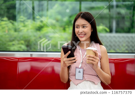 A charming Asian businesswoman is sipping coffee and reading messaged on her phone on the sky train. 116757370