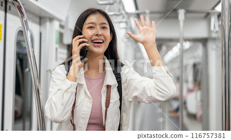 A cheerful Asian female is talking on the phone and greeting someone while commuting by sky train. A cheerful Asian female is talking on the phone and greeting someone while commuting by sky train. 116757381
