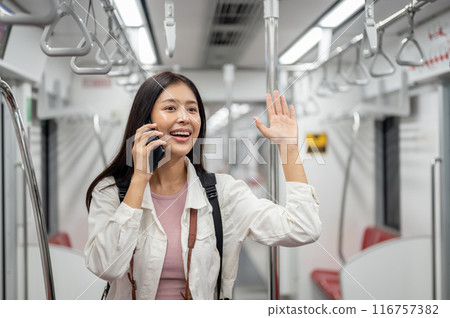 A cheerful Asian female is talking on the phone and greeting someone while commuting by sky train. A cheerful Asian female is talking on the phone and greeting someone while commuting by sky train. 116757382