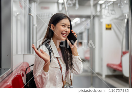 A happy Asian female tourist is talking on the phone while taking a sky train or metro train. 116757385