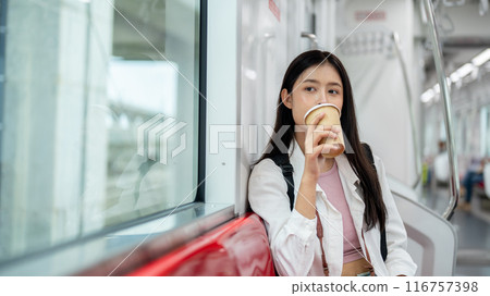 A happy young Asian female tourist is sipping coffee while sitting on a seat in a sky train. 116757398