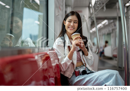 A happy young Asian female tourist is sipping coffee while sitting on a seat in a sky train. 116757400