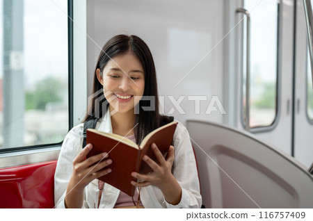 A happy Asian woman with a backpack is enjoying reading a book while taking a sky train somewhere. A happy Asian woman with a backpack is enjoying reading a book while taking a sky train somewhere. 116757409