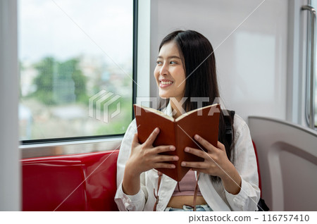 A happy Asian woman is looking at the view outside and reading a book while taking a sky train. A happy Asian woman is looking at the view outside and reading a book while taking a sky train. 116757410