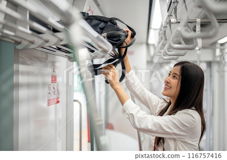 A happy female is putting her backpack in an overhead luggage space while commuting on a sky train. 116757416