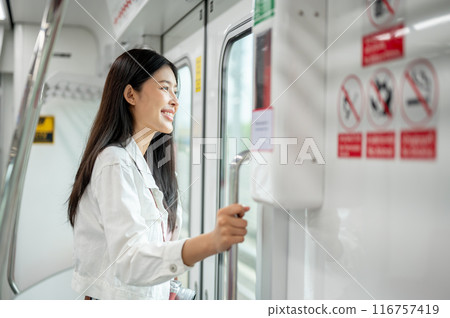A positive Asian female is looking at the view outside while holding onto a stanchion in a sky train 116757419