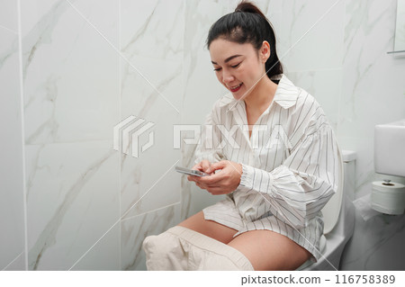 woman using smartphone and sitting on toilet bowl in bathroom 116758389