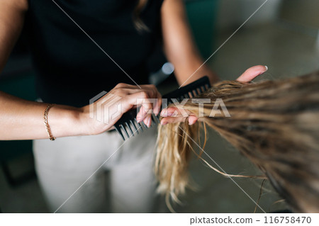 Closeup cropped shot of unrecognizable barber brushing wet long hair with hairbrush after washing. Close-up of blonde female client getting hair dressed in beauty salon. Concept of fashion hairstyling 116758470