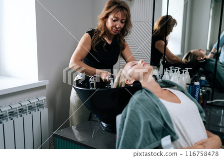 Female hairdresser carefully washing hair of beautiful blonde woman in beauty salon. Beautiful customer lady lying on salon washing bed feeling relax and happy enjoy massaging head. Female hairdresser carefully washing hair of beautiful blonde woman in beauty salon. Beautiful customer lady lying on salon washing bed feeling relax and happy enjoy massaging head. 116758478