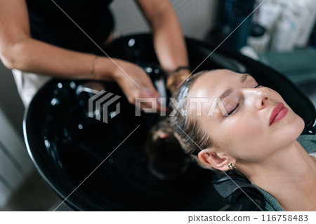 High-angle view of hairdresser washing head of young woman in beauty hairdressing salon. Close-up of unrecognizable hairstylist foaming shampoo on long blonde hair of beautiful female client. 116758483