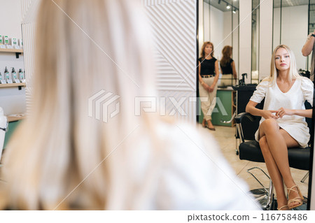 Over shoulder shot of young woman with long blonde hair patiently waiting for haircut sitting on chair in salon with casual and relaxed atmosphere. Concept of professional fashion hairstyling. Over shoulder shot of young woman with long blonde hair patiently waiting for haircut sitting on chair in salon with casual and relaxed atmosphere. Concept of professional fashion hairstyling. 116758486