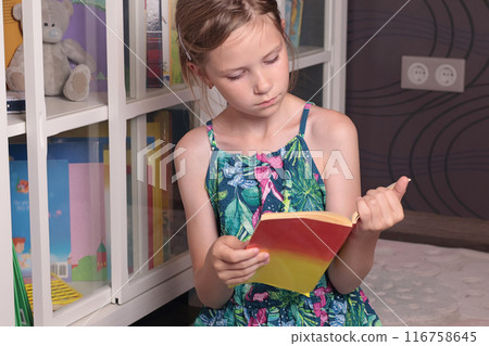 Teenage girl picks up, reads book from glass bookcase. Learning and reading. Hobbies and education.  116758645