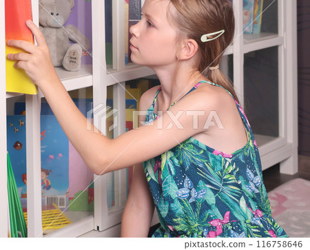 Teenage girl picks up, reads book from glass bookcase. Learning and reading. Hobbies and education.  116758646