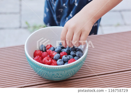 Child hand pick berries from bowl. Blueberries and raspberries. Family and lifestyle. Backyard in summer and nature, terrace.  116758729