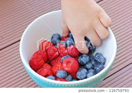 Child hand pick berries from bowl. Blueberries and raspberries. Family and lifestyle. Backyard in summer and nature, terrace. Child hand pick berries from bowl. Blueberries and raspberries. Family and lifestyle. Backyard in summer and nature, terrace. 116758793