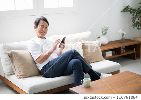 A man in his 50s sitting on a sofa and operating a smartphone 116761864