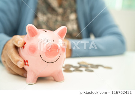 Asian elderly woman putting coin into pink piggy bank for saving money and insurance, poverty, financial problem in retirement. 116762431