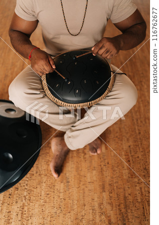 Top view vertical shot of unrecognizable spiritual African man in trance sitting playing on steel tongue drum on floor at home with cozy interior. Concept of mindfulness, tranquility, harmony. 116762677