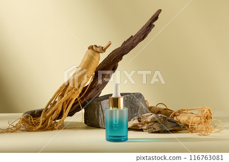 Front view photography with a blank label serum bottle in blue with gradient effect. In the back of it is some fresh ginseng roots, a log and stones, blank space for advertising 116763081
