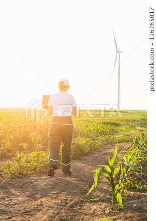 Woman farmer wearing white cap and t-shirt with laptop on agricultural field at sunset. Wind turbines in the background. Woman farmer wearing white cap and t-shirt with laptop on agricultural field at sunset. Wind turbines in the background. 116765017