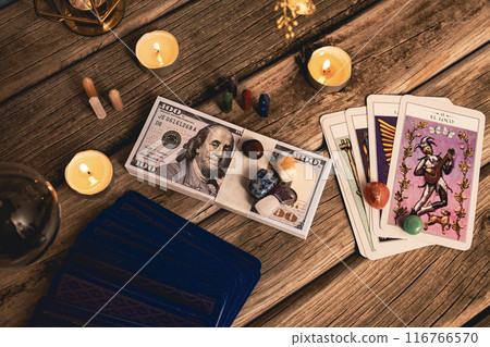 A tarot spread with The Fool card, hundred-dollar bills, and various crystals on a rustic wooden background with candles. 116766570