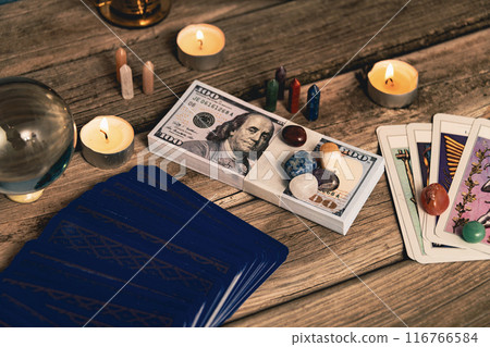 A tarot spread with The Fool card, hundred-dollar bills, and various crystals on a rustic wooden background with candles. 116766584