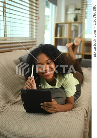 Carefree young Afro American woman in casual clothes lying on sofa and using a digital tablet 116766896