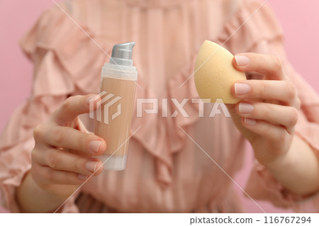 Female with bottle of foundation and cosmetic sponge in hands on pink background, close up 116767294
