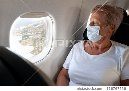 An elderly woman travels. Flying in a plane wearing a mask. Passenger. Watch the porthole. Look at the ground from above. Active lifestyle. An elderly woman travels. Flying in a plane wearing a mask. Passenger. Watch the porthole. Look at the ground from above. Active lifestyle. 116767536