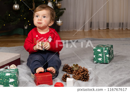 A one-year-old kid in a Santa hat opens gifts near the Christmas tree 116767617