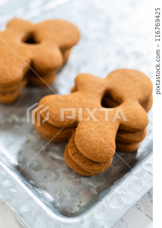 Gingerbread cookies, including a gingerbread man with a heart-shaped cutout, rest on a rustic metal tray against a marble countertop. 116769425