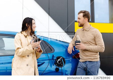Young couple man and woman traveling by electric car having stop at charging station. Boyfriend plugging in cable to charge. Man talking with girlfriend, holding cup drinking hot coffee smiling 116769490