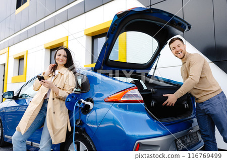 Young couple man and woman traveling by electric car having stop at charging station. The man taking a suitcase from the trunk. Man talking with girlfriend and she holding mobile phone in hand Young couple man and woman traveling by electric car having stop at charging station. The man taking a suitcase from the trunk. Man talking with girlfriend and she holding mobile phone in hand 116769499