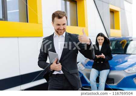 Young handsome car dealership consultant holding tablet while standing near car and showing finger up gesture Young handsome car dealership consultant holding tablet while standing near car and showing finger up gesture 116769504