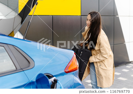 A young woman stands next to an electric car while charging the car at the station and puts her luggage in the trunk 116769770