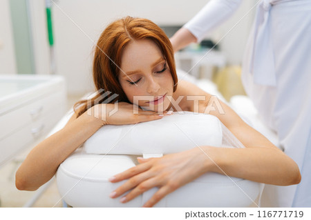 Closeup portrait of calm woman getting moxibustion treatment on back lying on massage table with closed eyes. Female client lying on spa bed during moxa smoke therapy. Concept of alternative medicine. Closeup portrait of calm woman getting moxibustion treatment on back lying on massage table with closed eyes. Female client lying on spa bed during moxa smoke therapy. Concept of alternative medicine. 116771719