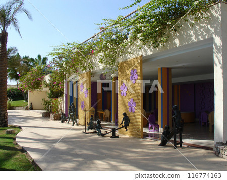Tropical resort building with decorative facade, large purple flowers, children sculptures, and lush landscaping. Welcoming entrance with palm trees. Tropical resort building with decorative facade, large purple flowers, children sculptures, and lush landscaping. Welcoming entrance with palm trees. 116774163
