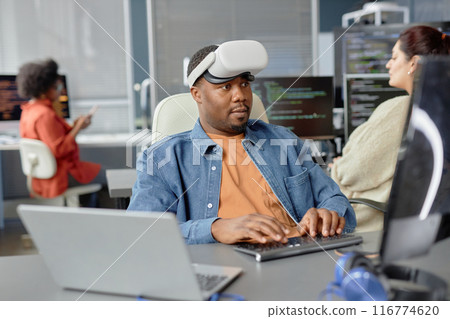 Portrait shot of software engineer of Black ethnicity in VR headset writing code for cutting-edge game development at working desk in office, copy space Portrait shot of software engineer of Black ethnicity in VR headset writing code for cutting-edge game development at working desk in office, copy space 116774620