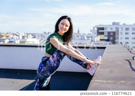 Beautiful brunette sexy woman in stylish sportswear stretching legs on the rooftop with beautiful city view on the sunset. After the workout. Beautiful brunette sexy woman in stylish sportswear stretching legs on the rooftop with beautiful city view on the sunset. After the workout. 116774621