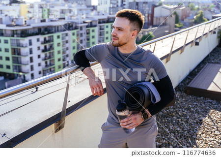 happy bearded sporty handsome man admiring city view on rooftop holding mat and bottle of water in his hands. Fitness male enjoy new goal concept. happy bearded sporty handsome man admiring city view on rooftop holding mat and bottle of water in his hands. Fitness male enjoy new goal concept. 116774636