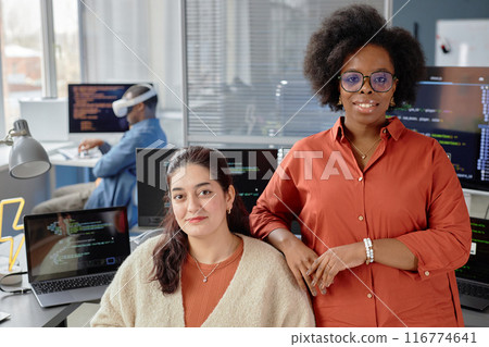 Portrait shot of two female multiethnic VR developers at working desk with computers looking at camera and smiling in IT company office Portrait shot of two female multiethnic VR developers at working desk with computers looking at camera and smiling in IT company office 116774641