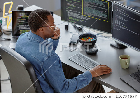 High-angle side view of thoughtful male African American software developer reading computer screen finding errors in code while sitting at desk with tea and candy bowl at hand in office, copy space 116774657